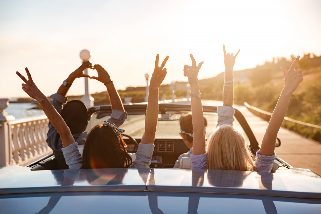 four friends in a top-down car with their hands raised as the sun shines on them while driving on a bridge near the ocean